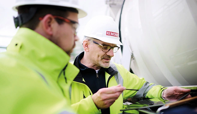 Two REMONDIS employees wearing high-visibility jackets and white helmets, engaged in conversation while examining equipment and looking at a tablet.