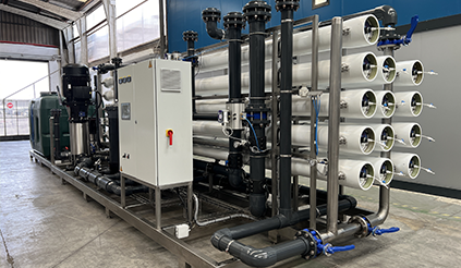 Industrial reverse osmosis water filtration system, featuring multiple horizontal white cylindrical membrane housings mounted on a metal frame with black piping and valves. The system includes a control panel and pumps and is situated inside a large industrial facility.